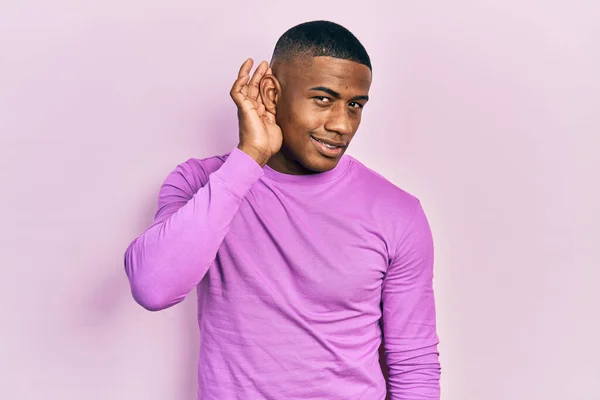 噂やゴシップに耳を傾ける耳の上に手で笑みを浮かべてカジュアルなピンクのセーターを着て若い黒人男性 聴覚障害の概念 — ストック写真