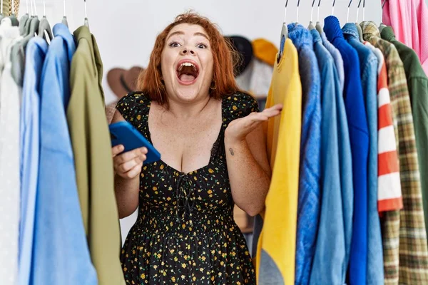 Young Redhead Woman Searching Clothes Clothing Rack Using Smartphone Celebrating — Stockfoto