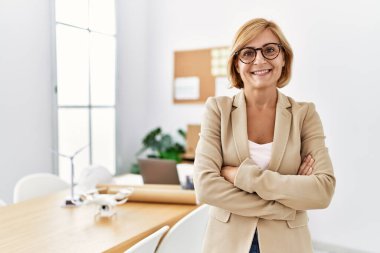 Orta yaşlı sarışın kadın, mimarlık stüdyosunda kollarını kavuşturup gülümsüyor.