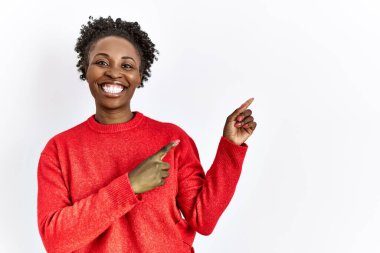 Genç Afro-Amerikalı kadın izole edilmiş arka planda günlük giysiler giyiyor gülümsüyor ve kameraya bakıyor iki eliyle ve parmaklarıyla yan tarafa bakıyor.. 