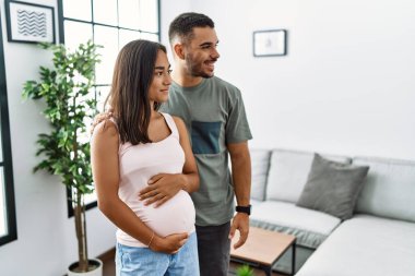 Bir bebek bekleyen ırklar arası genç bir çift, hamile karnına dokunup yan tarafa bakıyor, rahat bir görünüm takınıyor ve kendine güvenen bir gülümsemeyle gülümsüyor.. 