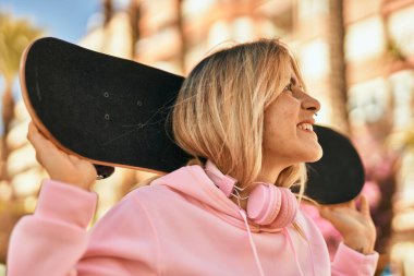 Genç sarışın kaykaycı kız gülümseyerek şehirde kaykay yapıyor..