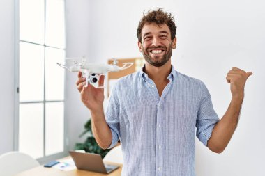 Young handsome man using drone at architect office pointing thumb up to the side smiling happy with open mouth 