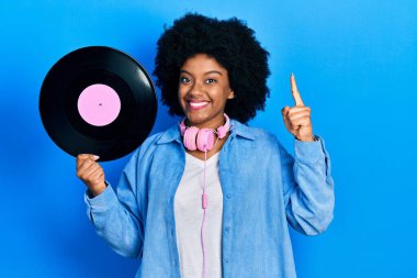 Kulaklık takan, elinde vinil diskle gülümseyen, mutlu yüzlü, soru işaretli genç Afrikalı Amerikalı bir kadın. 