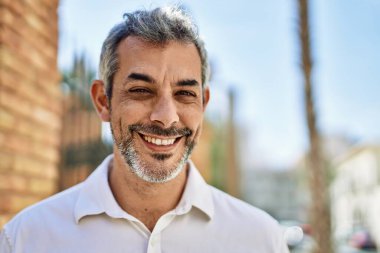 Orta yaşlı, kır saçlı, gülümseyen bir adam şehirde mutlu bir şekilde duruyor..