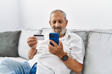 Kıdemli gri saçlı adam evde akıllı telefon ve kredi kartı kullanarak gülümsüyor.