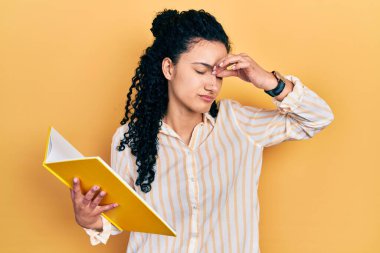Young hispanic woman with curly hair holding book tired rubbing nose and eyes feeling fatigue and headache. stress and frustration concept. 