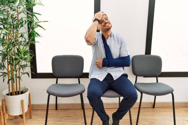 Yakışıklı genç adam, doktor bekleme odasında kolundan sakatlanmış, gülümsüyor, saçlarına dokunuyor, el hareketi yapıyor, çekici ve şık duruyor. 