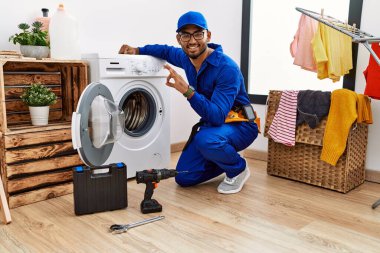 Çamaşır makinesinde çalışan genç Hintli teknisyen gülümseyip el ve parmaklarıyla 