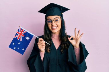 Mezuniyet üniforması giyen, elinde Avustralya bayrağı olan genç İspanyol kadın parmaklarıyla işaret veriyor, gülümseyerek mükemmel bir sembol oluşturuyor. 