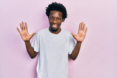Genç Afro-Amerikan adam, günlük beyaz tişört giyer ve parmaklarıyla on numarayı gösterirken kendinden emin ve mutlu bir şekilde gülümser.. 