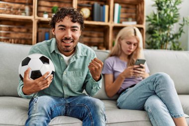 Adam mutlu bir şekilde futbol maçı izliyor ve kız arkadaşı evde akıllı telefon kullanıyor..