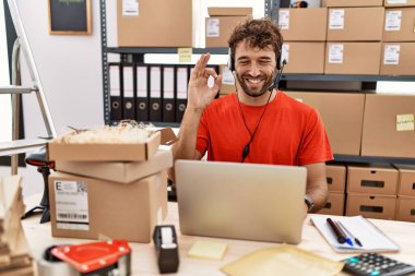 Young hispanic call center agent man working at warehouse smiling positive doing ok sign with hand and fingers. successful expression. 