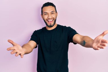 Siyah bir tişört giyen İspanyol sakallı adam kameraya sarılıp gülümsüyor. Mutluluğu kucaklayan neşeli bir ifade.. 