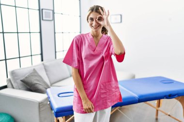 Young blonde woman working as physiotherapist at home doing ok gesture with hand smiling, eye looking through fingers with happy face. 