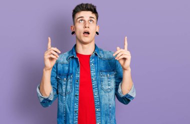 Kulakları genişlemiş, sıradan bir kot ceket giymiş genç beyaz çocuk şaşırmış ve şaşırmış bir şekilde yukarı bakıyor ve parmaklarıyla işaret ediyor ve kollarını kaldırıyor.. 