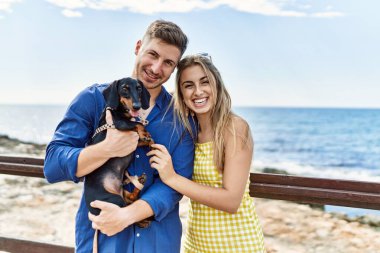 Kumsalda mutlu gülümseyen ve köpekle kucaklaşan beyaz bir çift..