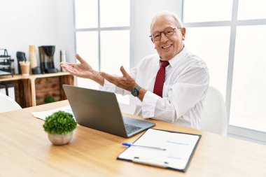 Ofiste çalışan kıdemli bir adam bilgisayarı kullanıyor. İçeri girmeye davet ediyor. Doğal bir şekilde gülümsüyor. 