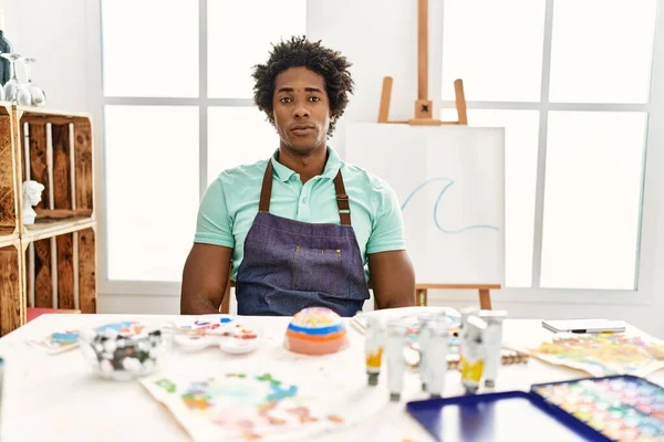 年轻的非洲裔美国人坐在艺术工作室的桌子上 脸上带着严肃的表情 简单而自然地看着相机 — 图库照片