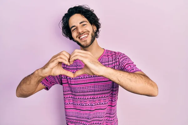 手でハートのシンボルの形をして愛に笑みを浮かべてカジュアルなピンクのTシャツを着てハンサムなヒスパニック系の男 ロマンチックなコンセプト — ストック写真