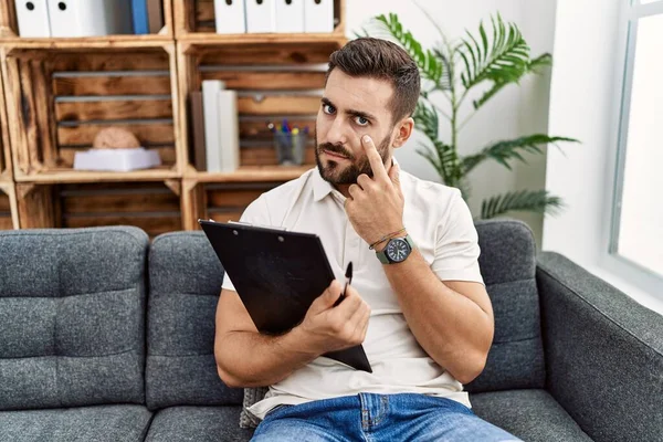 Handsome Hispanic Man Holding Clipboard Working Psychology Clinic Pointing Eye — Foto de Stock
