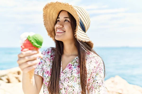 年轻的拉丁女孩戴着夏帽 坐在海滩的岩石上吃着冰激凌 — 图库照片