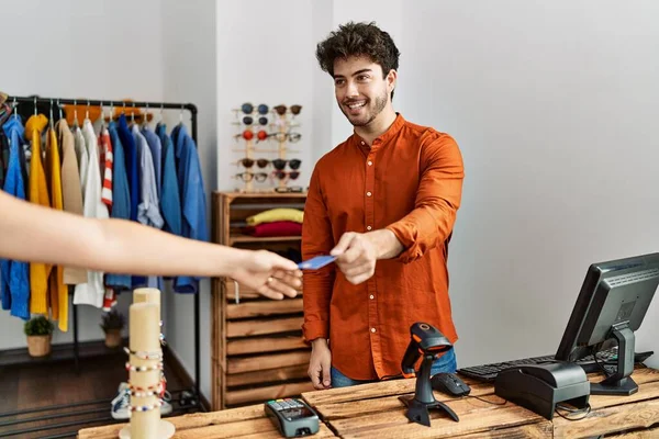 Young hispanic shopkeeper man giving credit card to customer at clothing store.