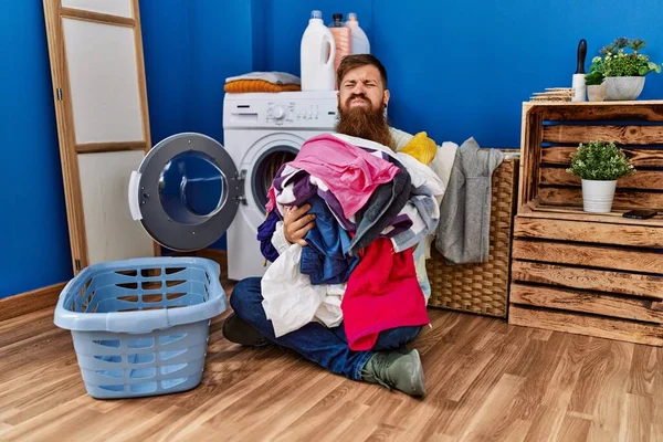 红头发的男人留着长胡子 把脏衣服放进洗衣机里 脸上带着滑稽的表情 嘴被空气吹胀 — 图库照片