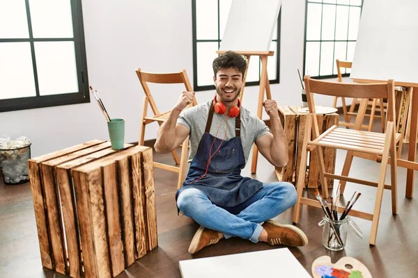 Young hispanic man sitting at art studio excited for success with arms raised and eyes closed celebrating victory smiling. winner concept. 