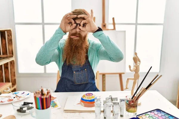 红头发男人 留着长胡子 在艺术工作室里画着黏土碗 做着像双筒望远镜一样的手势 伸出舌头 眼睛透过手指看着 疯狂的表达 — 图库照片