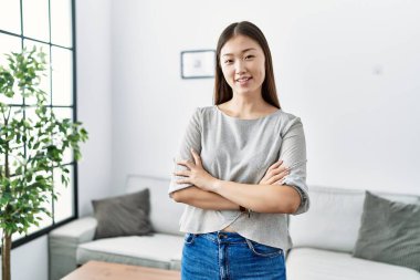 Genç Asyalı kadın oturma odasında kollarını kavuşturup gülümsüyor.
