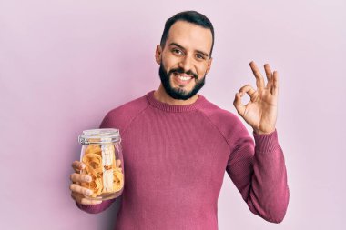 Sakallı, elinde makarna kavanozu olan genç adam parmaklarıyla imza atıyor, gülümseyerek mükemmel bir sembol oluşturuyor. 