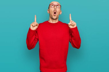 Bald man with beard wearing casual clothes and glasses amazed and surprised looking up and pointing with fingers and raised arms. 