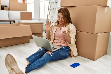 Orta yaşlı, beyaz bir kadın yeni evinde yerde otururken bilgisayarla video görüşmesi yapıyor..