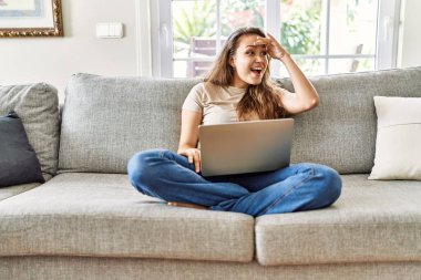 Güzel, genç, esmer bir kadın evdeki bilgisayar laptopunu kullanarak koltukta oturuyor. Çok mutlu ve gülümsüyor, başını kaldırıp uzaklara bakıyor. arama kavramı. 