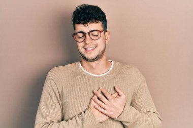 Gündelik kıyafetler ve gözlük takan genç İspanyol adam elleri göğsünde, gözleri kapalı ve yüzünde minnet dolu bir jest ile gülümsüyor. sağlık kavramı. 