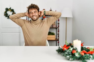 Young handsome man with beard sitting on the table by christmas decoration relaxing and stretching, arms and hands behind head and neck smiling happy 
