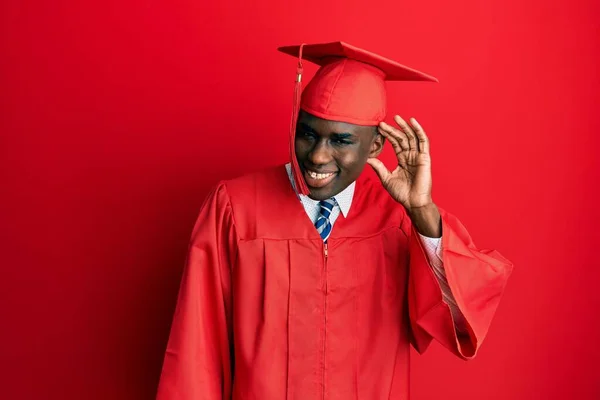 卒業キャップと儀式用のローブを身に着けている若いアフリカ系アメリカ人の男性は 噂やゴシップに耳を傾け手で笑みを浮かべています 聴覚障害の概念 — ストック写真
