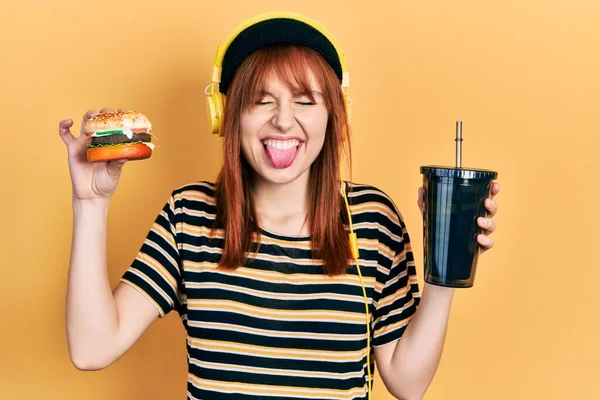 红头发的年轻女人吃汉堡包 喝苏打水 戴着耳机 带着滑稽的表情伸出舌头 高兴极了 — 图库照片