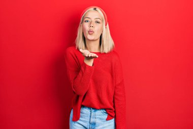 Güzel sarışın kadın, gündelik kırmızı kazak giyip kameraya bakıyor ve havada bir öpücük savuruyor. Güzel ve seksi. aşk ifadesi. 