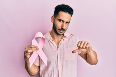 Öfkeli yüzlü pembe kanser kurdelesi tutan genç İspanyol adam, olumsuz işaret baş parmakları aşağıda, reddedilme konsepti. 