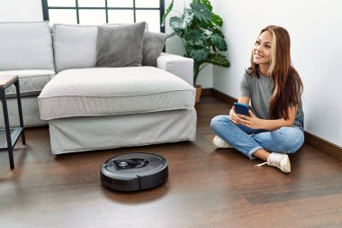 Evde yerde oturan akıllı telefonuyla robot elektrik süpürgesini kontrol eden beyaz bir kız..