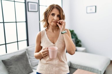 Orta yaşlı kadın evde kahve içiyor ve dudakları parmaklarıyla fermuar gibi kapalı. gizli ve sessiz, tabu konuşması 