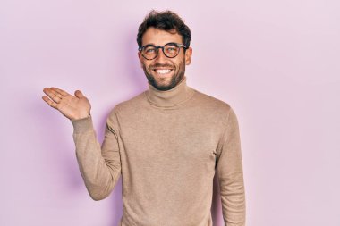 Handsome man with beard wearing turtleneck sweater and glasses smiling cheerful presenting and pointing with palm of hand looking at the camera. 