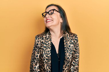 Orta yaşlı İspanyol bir kadın iş kıyafetleri ve gözlük takıyor. Yüzünde gülümseme, doğal bir ifade ile yan yana bakıyor. Kendinden emin gülen. 