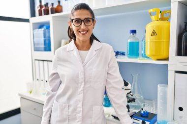 Laboratuvarda dikilen, bilim adamı üniforması giyen genç İspanyol kadın.