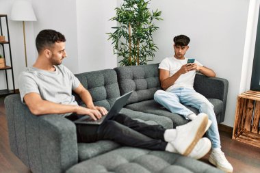 Two hispanic men couple on disagreement using smartphone and laptop at home