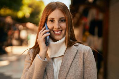 Genç kızıl saçlı kız gülümsüyor. Şehirdeki akıllı telefondan konuşuyor..