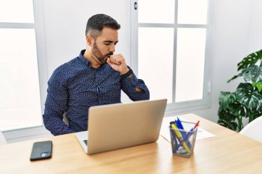 Ofiste sakallı, laptopla çalışan, kendini kötü hisseden ve soğuk algınlığı veya bronşitin belirtisi olarak öksüren genç İspanyol bir adam. sağlık hizmeti kavramı. 