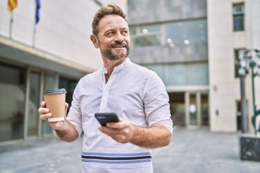 Şehirde akıllı telefon kullanan orta yaşlı bir adam kahve tutuyor.